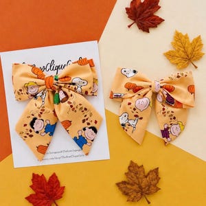 May include: Two light orange fabric hair bows featuring a repeating pattern of Peanuts characters including Snoopy, Charlie Brown, and Lucy, along with pumpkins and autumn leaves. The bows are displayed on a white card with black text.