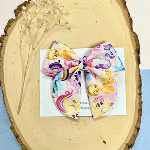 May include: A fabric hair bow featuring a repeating pattern of colorful cartoon ponies on a light pink background. The bow is centered on a white rectangular display card with partial black text visible, including the word BowClips.