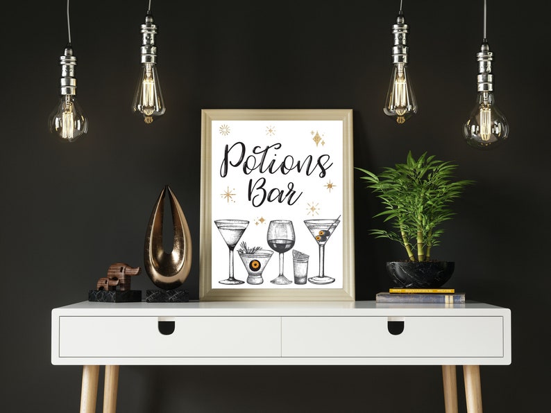 May include: A framed "Potions Bar" print with illustrations of cocktails. The print is displayed on a white table with decorative items, including a gold sculpture, a small elephant statue, and a potted plant. Overhead, there are hanging light fixtures.