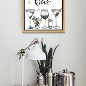 May include: A framed print with the words "Potions Bar" and illustrations of cocktail glasses. The print is displayed above a bar cart with a white lamp, cocktail shakers, and glassware. The overall theme is a Halloween or cocktail party.