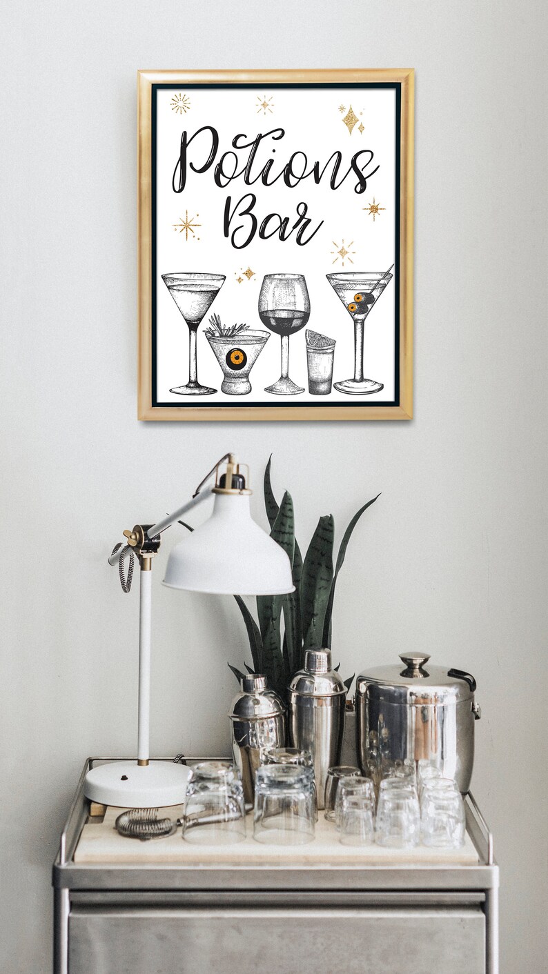 May include: A framed print with the words "Potions Bar" and illustrations of cocktail glasses. The print is displayed above a bar cart with a white lamp, cocktail shakers, and glassware. The overall theme is a Halloween or cocktail party.