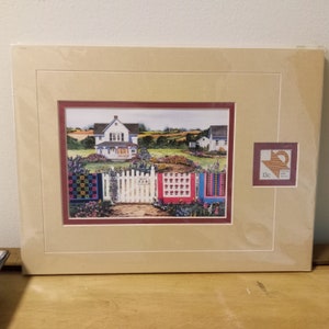 May include: A framed print of a watercolor painting depicting a white farmhouse with a picket fence and colorful quilt squares. The print includes a vintage postage stamp with a 13c denomination.