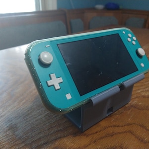 May include: A teal Nintendo Switch Lite handheld gaming console in a clear protective case, sitting on a gray stand on a wooden table.