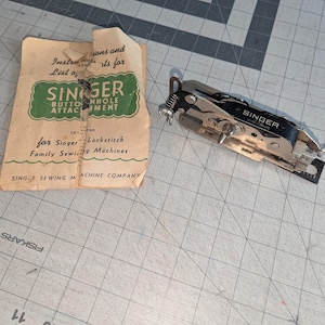 May include: A vintage Singer buttonhole attachment for Singer Lockstitch family sewing machines. The attachment is silver with a black Singer label. The attachment is shown with its original instructions.