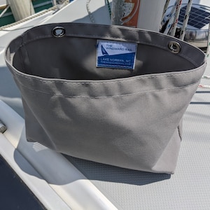 May include: A gray canvas storage bag with a rectangular shape and a wide opening. The bag has metal grommets at the top and a label that reads "THE WINDWARD RAIL LAKE NORMAN, NC". The bag is designed for use on a boat.