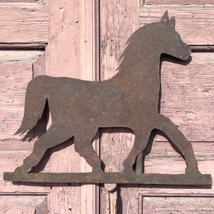 Pode incluir: Uma silhueta de cavalo de metal enferrujado, montada em uma superfície de madeira. O cavalo está voltado para a direita e tem uma cauda e uma crina longas.