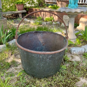 May include: A large, antique copper pot with a rusty iron handle. The pot is green with age and has a hammered texture. It is sitting on a bed of green grass.