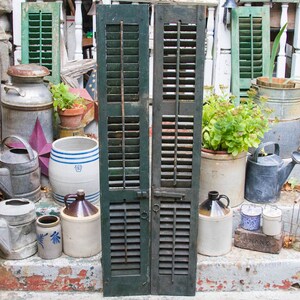 Puede incluir: Dos contraventanas de madera verde envejecidas con un pestillo de metal. Las contraventanas están en un conjunto de escalones de piedra con varios artículos vintage, incluyendo regaderas, jarras y una gran tina.