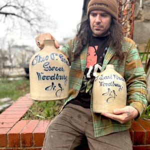 Puede incluir: Dos tinajas de gres vintage con letras azules. La tinaja más grande dice "Theo F. Green Grocer, Woodbury, N.J." y la tinaja más pequeña dice "J. Earl Budd, Woodbury, N.J."
