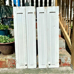 May include: Two white wooden shutters with vertical panels and a knob on each. The shutters are closed and are leaning against a set of stairs.