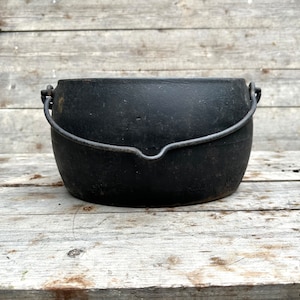 May include: A black cast iron cauldron with a curved metal handle. The cauldron is round with a wide opening and sits on a wooden surface. The metal handle is attached to the sides of the cauldron.