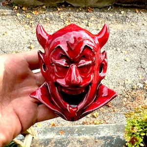 May include: A red ceramic devil head with horns and a wide, toothy grin. The devil head is glazed with a glossy finish.