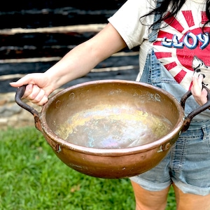 May include: A large, antique copper bowl with two curved, dark metal handles. The bowl has a wide, open design and a weathered, aged appearance. The bowl is held by a person wearing denim overalls and a graphic t-shirt.