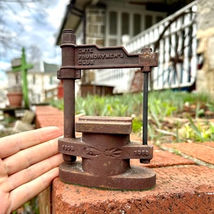 May include: A rusty metal sculpture of a small, hand-operated press. The sculpture is labeled "CMTE FOUNDRYMEN'S CLUB" and has the dates "1908" and "1968" on it.