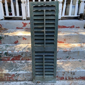 May include: A weathered green wooden window shutter with multiple louvers. The shutter is closed and has a distressed paint finish.