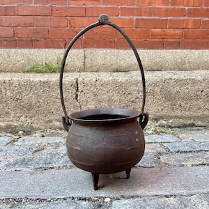 May include: A rustic, iron cauldron with a handle, standing on four legs. The cauldron is weathered and has a dark brown patina.