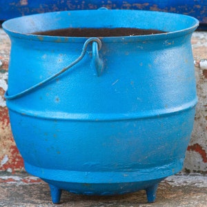 May include: A blue cast iron pot with a handle. The pot has a worn, rustic look and is sitting on a concrete surface.