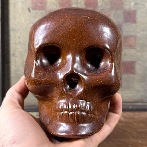 May include: A brown skull sculpture with a glossy finish. The skull has detailed features, including eye sockets, a nasal cavity, and teeth. The sculpture is held in a hand, showcasing its size and shape. The skull is a decorative item.