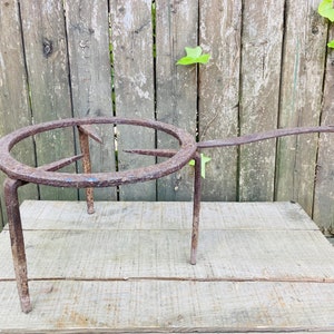 May include: A rusty metal tripod stand with a circular top. The stand is made of thick metal and has a rustic appearance.
