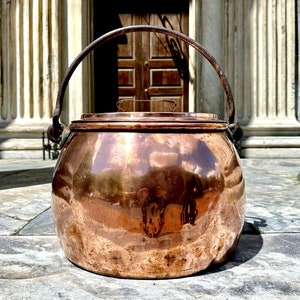 May include: A large, antique copper pot with a lid and a handle. The pot has a rounded body and a slightly flared rim. The handle is attached to the pot with two rivets. The pot is sitting on a stone surface.