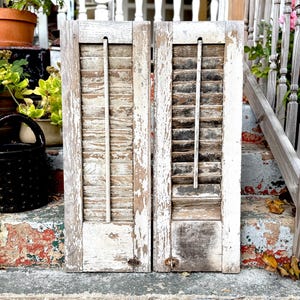 May include: Two weathered, white-painted wooden shutters with horizontal slats and a central vertical support. The shutters show signs of age with peeling paint and a distressed appearance, suggesting a vintage or rustic aesthetic. The shutters are leaning against a staircase.