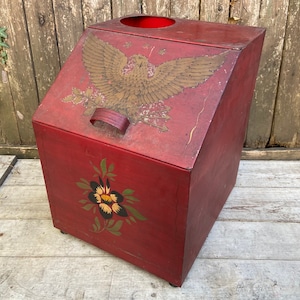May include: Red metal storage bin with a painted design of a golden eagle and a flower. The bin has a hinged lid and a handle.