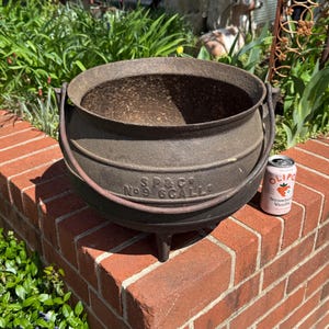 May include: A large, cast iron cauldron with two handles and a worn, rustic finish. The cauldron is sitting on a brick wall and has the text "SP&Co No 6 GALLS" embossed on the side.