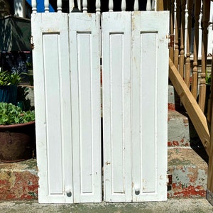 Puede incluir: Un par de contraventanas de madera blanca con paneles verticales y una simple perilla en cada puerta. Las contraventanas están cerradas y parecen viejas y desgastadas.