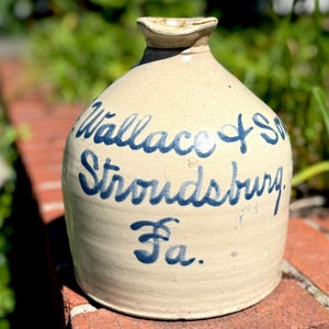 Puede incluir: Una jarra de gres vintage con un esmalte beige. La jarra presenta letras azules en cursiva que dicen "Wallace + Son Stroudsburg Pa." La jarra tiene un cuerpo redondeado y un pequeño pico. Está sobre una superficie de ladrillo rojo.