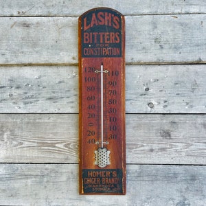 May include: A vintage wooden thermometer with a red and black sign advertising "Lash's Bitters for Constipation" and "Homer's Chiger Brandy for Diarrhea Stomach Ache".