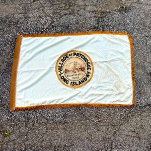 May include: A white flag with a gold fringe border. The flag features a circular seal with the text "Village of Patchogue, Long Island, NY" and "Incorporated 1898".