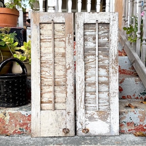 May include: Two weathered, vintage wooden shutters with peeling white paint. Each shutter has vertical slats and a central vertical support. The shutters are propped against a brick and concrete surface, suggesting outdoor use or decor.