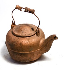May include: A vintage copper-colored cast iron teapot with a rounded body, spout, and a hinged lid. The handle is made of a curved metal with a decorative, textured grip. The teapot is likely used for brewing tea.