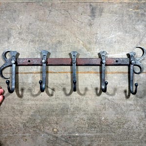 Puede incluir: Un perchero de metal rústico con cinco ganchos. El perchero está hecho de metal oscuro con un acabado marrón. Los ganchos tienen forma de hojas estilizadas y tienen una textura martillada.