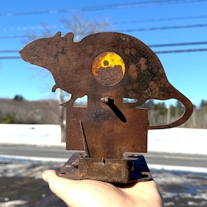 May include: A rusty metal rat silhouette target with a yellow bullseye. The target is mounted on a metal base with a lever.