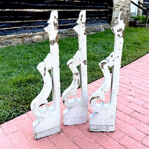 May include: Three antique white wooden corbels with distressed paint. The corbels have a decorative scroll design and are standing on a pink brick surface. The background includes green grass and a rustic wooden building.