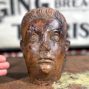 May include: A weathered, brown sculpture of a human head with detailed facial features. The sculpture has a textured surface and appears to be made of a composite material. The head is a classical bust style, with a focus on the eyes and mouth.