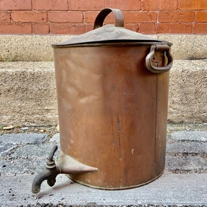 Może przedstawiać: Antyczny miedziany chłodnik do wody z pokrywką i kranem. Chłodnik jest wykonany z miedzi i ma patynowane wykończenie. Ma uchwyt na pokrywce i wylewkę z boku.