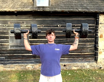 May include: A person holding two large, black dumbbells overhead. The person is wearing a blue t-shirt with the word "Colce" printed on the front and white shorts. The background is a wooden structure.