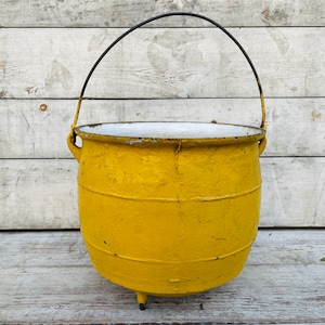 Puede incluir: Un caldero de hierro fundido amarillo con un asa de metal negro. El caldero está sentado sobre una superficie de madera blanca.