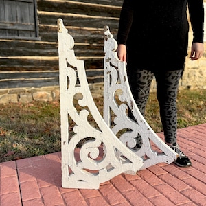 May include: Two white, ornate wooden corbels with scrollwork and a distressed finish. The corbels are triangular in shape and appear to be vintage or antique architectural elements. They are displayed outdoors on a brick surface.