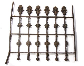 May include: A rusty metal fence panel with decorative ironwork. The fence has six vertical bars with ornate finials at the top and star-shaped accents in the middle. The fence is made of dark brown metal with signs of rust and wear.