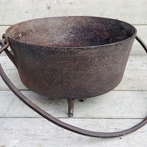 May include: A rustic cast iron cauldron with three legs and a single handle. The cauldron is made of dark metal and has a worn, aged appearance.
