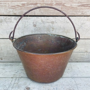 Puede incluir: Un cubo de cobre con un asa de alambre. El cubo está vacío y tiene un aspecto desgastado y envejecido.