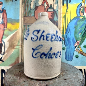 May include: A white ceramic jug with blue lettering that reads "M. Sheehan & Co.  " The jug has a rounded body and a wide mouth. It is sitting on a gray surface.