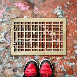 May include: A rusty metal vent cover with a square grid pattern, set against a red, brown, and white painted concrete floor. Two red sneakers are visible in the bottom of the image.