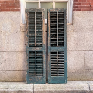 May include: Two weathered, teal-colored wooden shutters with louvered panels, leaning against a brick and stone wall.