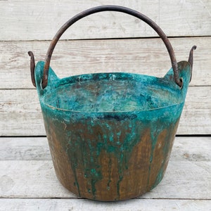 May include: A vintage copper bucket with a green patina and a rusty iron handle. The bucket is sitting on a white wooden surface.