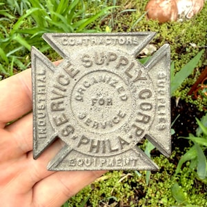 Puede incluir: Un emblema de metal en forma de cruz con el texto "Industrial Service Supply Corp. Organizado para el servicio - Phila. Equipo Contratistas Suministros" alrededor del círculo central.