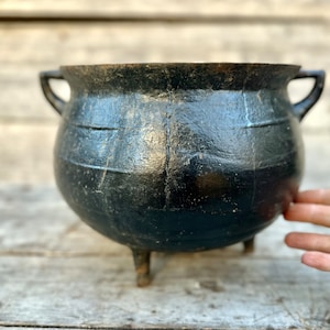 May include: A black cast iron cauldron with three legs and two handles. The cauldron is slightly worn and has a rustic appearance.